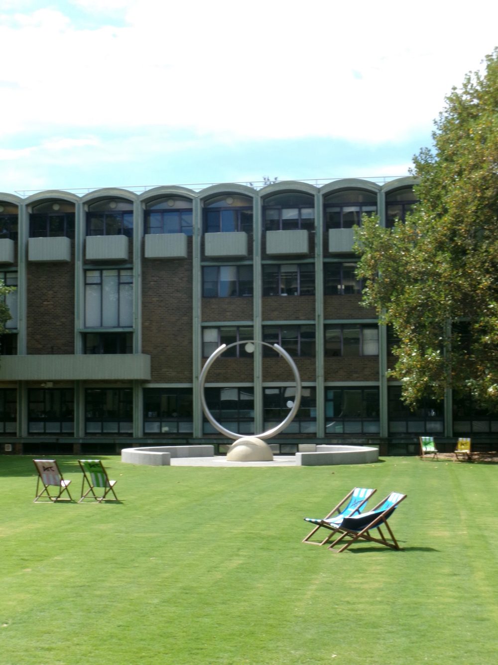 Deck chairs on a green lawn before a ring sculpture and a brick building